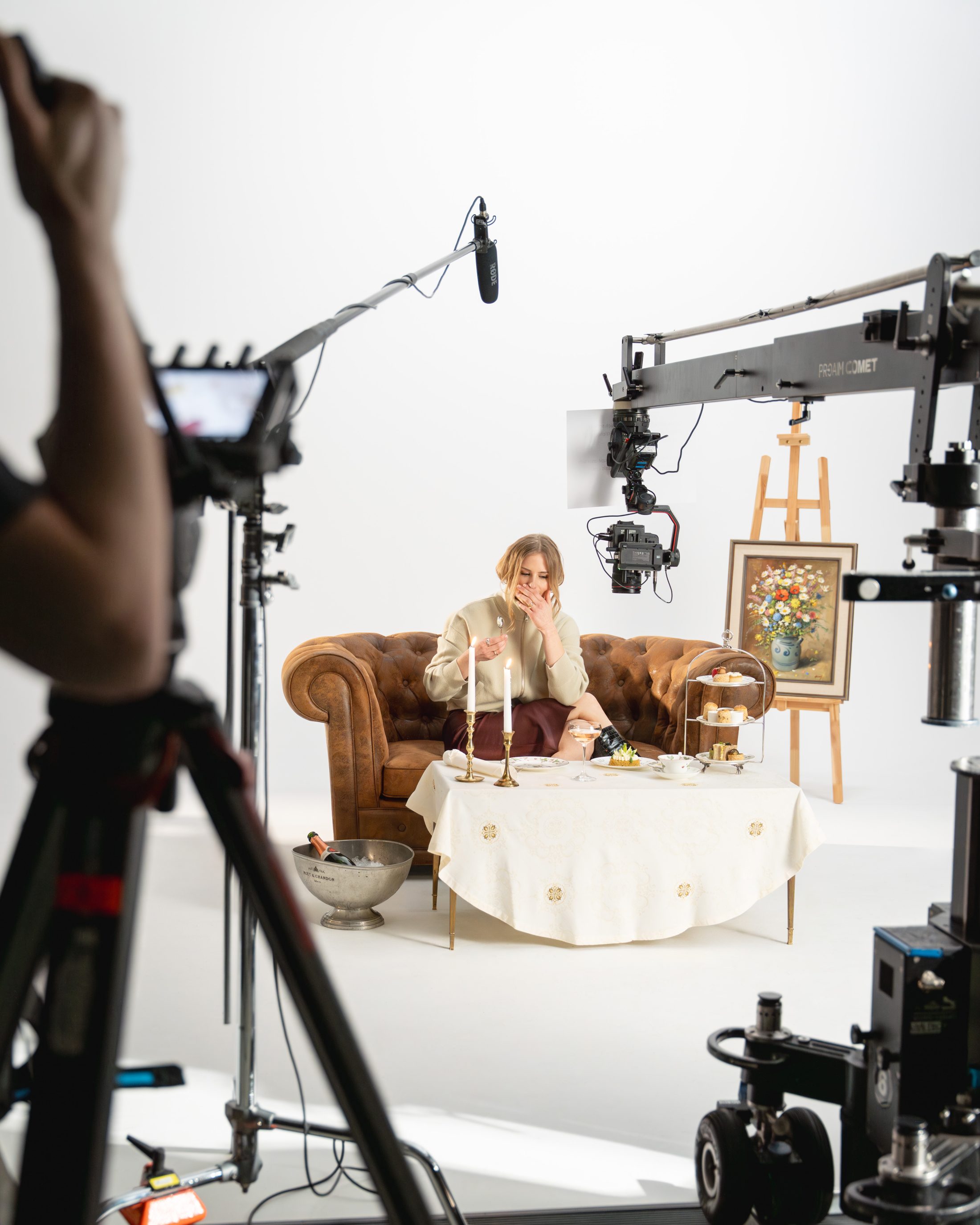 Blick hinter die Kulissen des Shootings von «SR-STUDIOS has great taste» in einem hellen Studio. Eine blonde Frau sitzt im Hintergrund auf einem braunen Ledersofa vor einem schick gedeckten Tisch. Im Vordergrund sind Kameras, Stative und Studiobeleuchtung zu sehen. | SR-STUDIOS