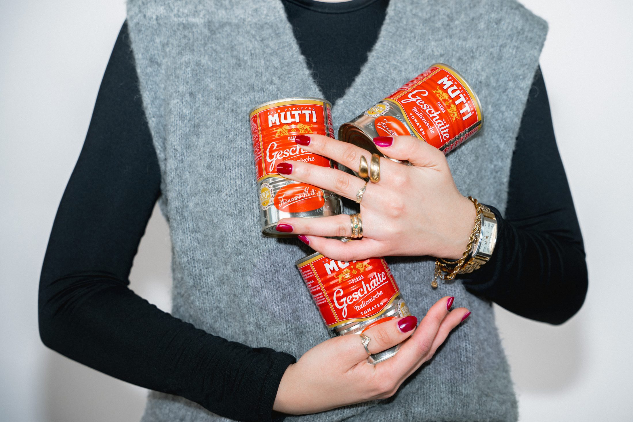 Lifestyle Fotografie und Brand Awareness: Eine Frau mit goldenem Schmuck und roten Fingernägeln präsentiert drei rote Mutti Tomatendosen als Teil eines Shootings. SR-STUDIOS. | SR-STUDIOS