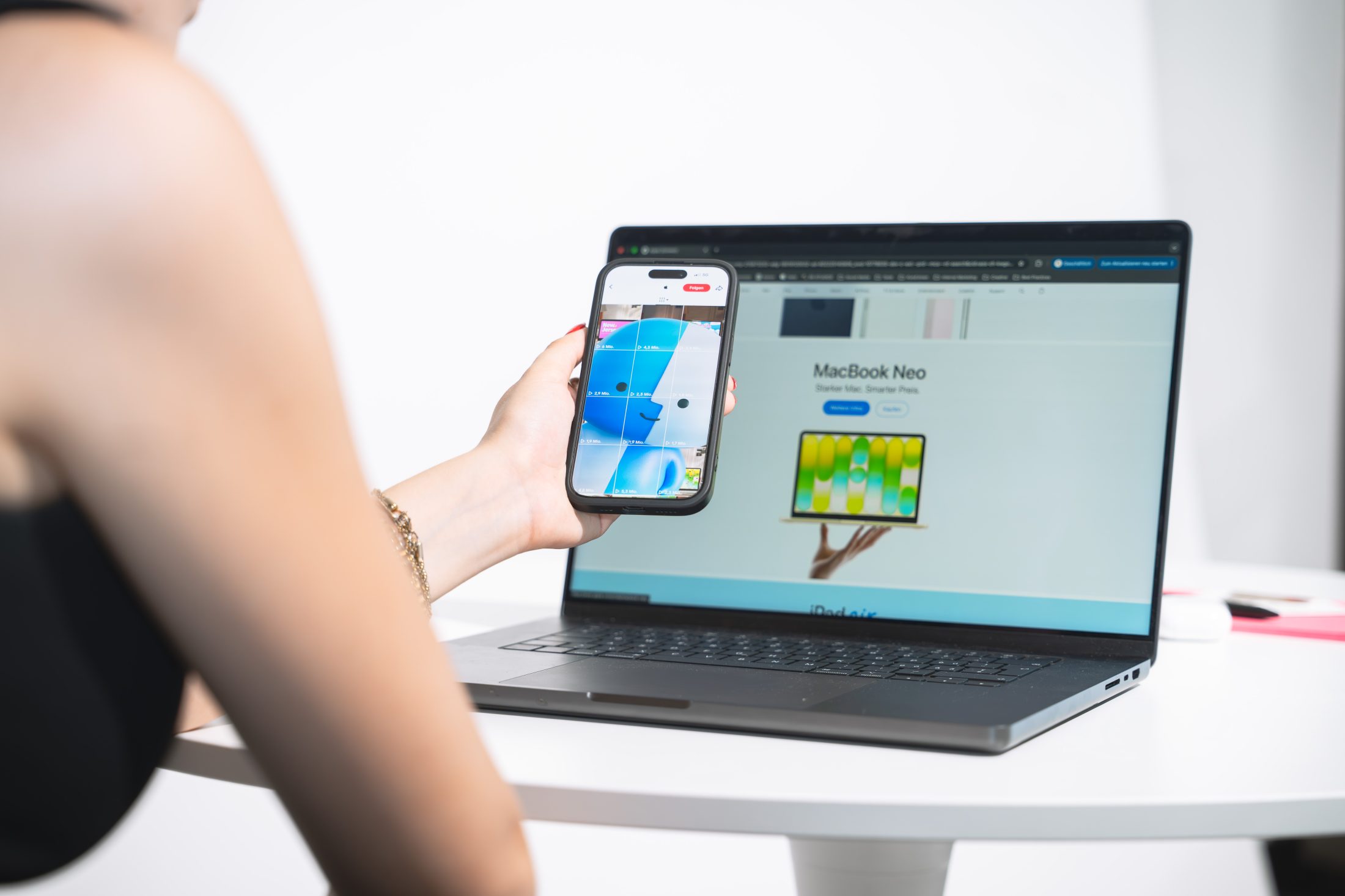 Nahaufnahme einer Frau, die ein Smartphone in der Hand hält, das das blaue Finder-Maskottchen (Lil' Finder Guy von Apple) im TikTok-Feed zeigt. Im Hintergrund steht ein offenes MacBook Neo auf einem weißen Tisch, auf dessen Display die Produktseite mit dem Slogan «MacBook Neo: Starker Mac. Smarter Preis.» zu sehen ist. Brand Maskottchen Chitchat für SR-STUDIOS. | SR-STUDIOS