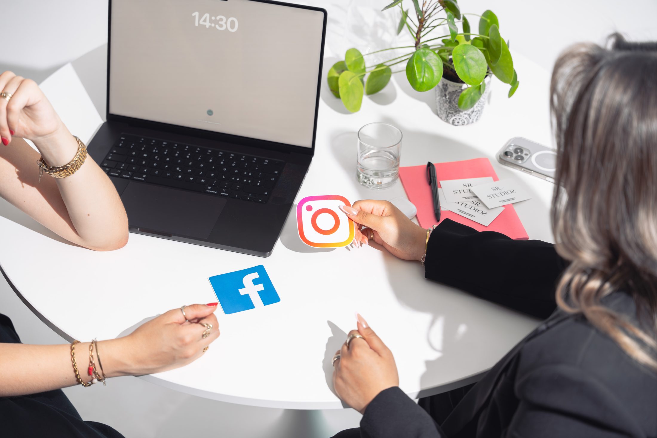Zwei Digital Marketing Specialist diskutieren Meta Advertising, sitzen an einem weissen Tisch mit Laptop und halten das Instagram und Facebook Logo hoch; SR-STUDIOS. | SR-STUDIOS