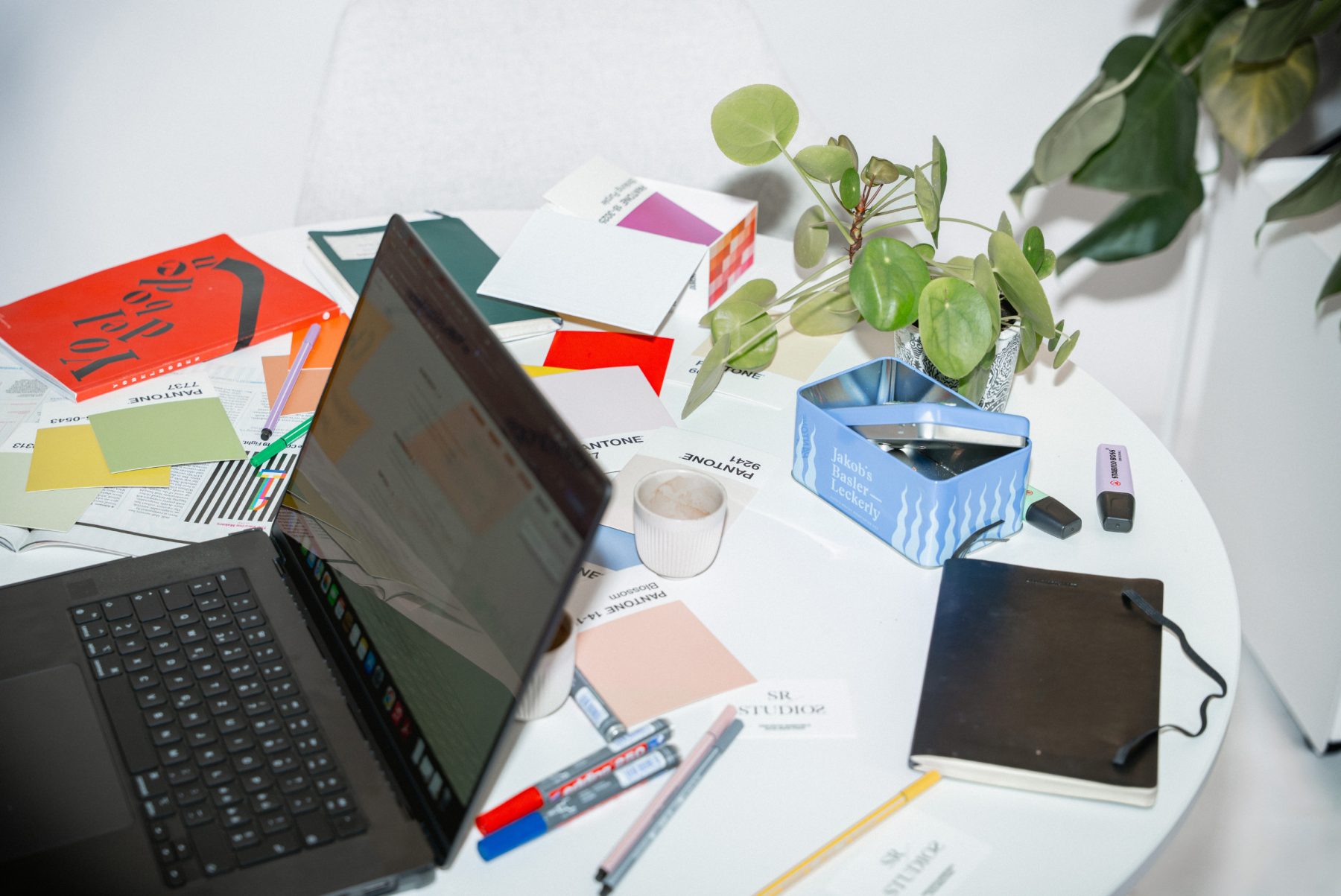 Überblick auf einen geschäftigen, kreativen Arbeitsplatz bei SR-STUDIOS. Zu sehen sind ein offener Laptop, Pantone-Farbfächer, Marker, Notizbücher, eine Dose «Jakob's Basler Leckerly» und Zimmerpflanzen auf einem runden weißen Tisch, umgeben von Designer-Stühlen. | SR-STUDIOS