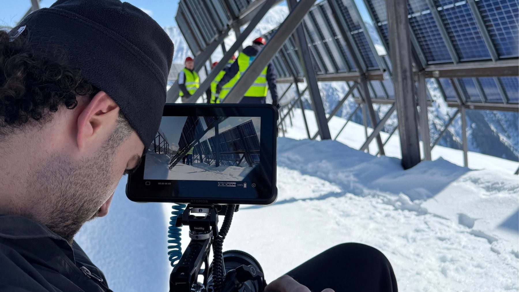 Nahaufnahme eines SR-STUDIOS Kameramanns (Content Creator) im Schnee, der die Komposition auf dem Monitor seiner Kamera kontrolliert. Im Hintergrund sind aufgestellte Solaranlagen der Madrisa Solar AG und Mitarbeiter:innen in Warnwesten und Schutzhelmen zu sehen. | SR-STUDIOS
