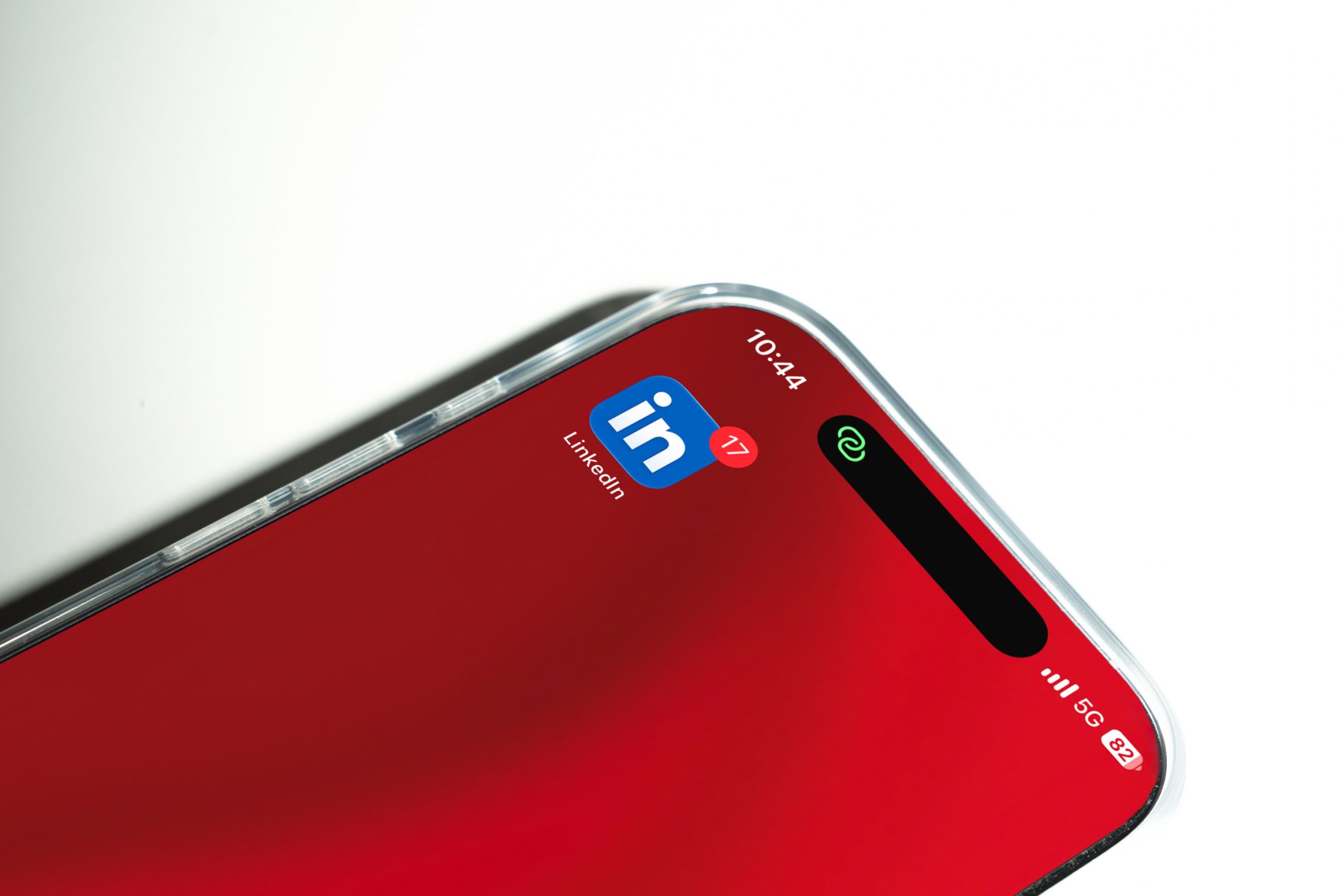 Ein Chitchat über das Marketingtool LinkedIn von SR-STUDIOS. Nahaufnahme der oberen Ecke eines iPhone / Smartphones mit transparentem Case, das auf einem weissen Untergrund liegt. Auf dem roten Bildschirmhintergrund ist das blaue LinkedIn-App-Icon mit einer roten Benachrichtigungszahl (17) zu sehen, daneben die Uhrzeit 10:44 sowie die Statusanzeigen für Empfang (5G) und Akku (82%). | SR-STUDIOS