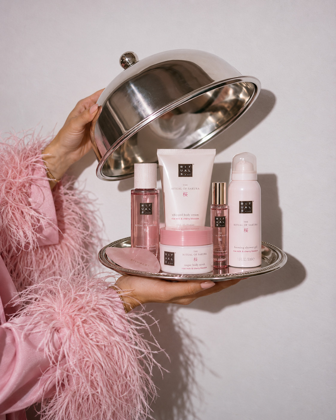 «Eine Chitchat Success Story über Rituals von SR-STUDIOS»: Auf einem silbernen Tablett werden diverse Pflegeprodukte der Reihe «The Ritual of Sakura» von Rituals präsentiert, darunter ein Körperpeeling, eine Körpercreme und ein Duschschaum. Ein rosa Guasha-Stein liegt ebenfalls auf dem Tablett. Eine Person in einem rosa, flauschigen Bademantel hält das Tablett, während eine andere Person eine silberne Cloche anhebt. | SR-STUDIOS