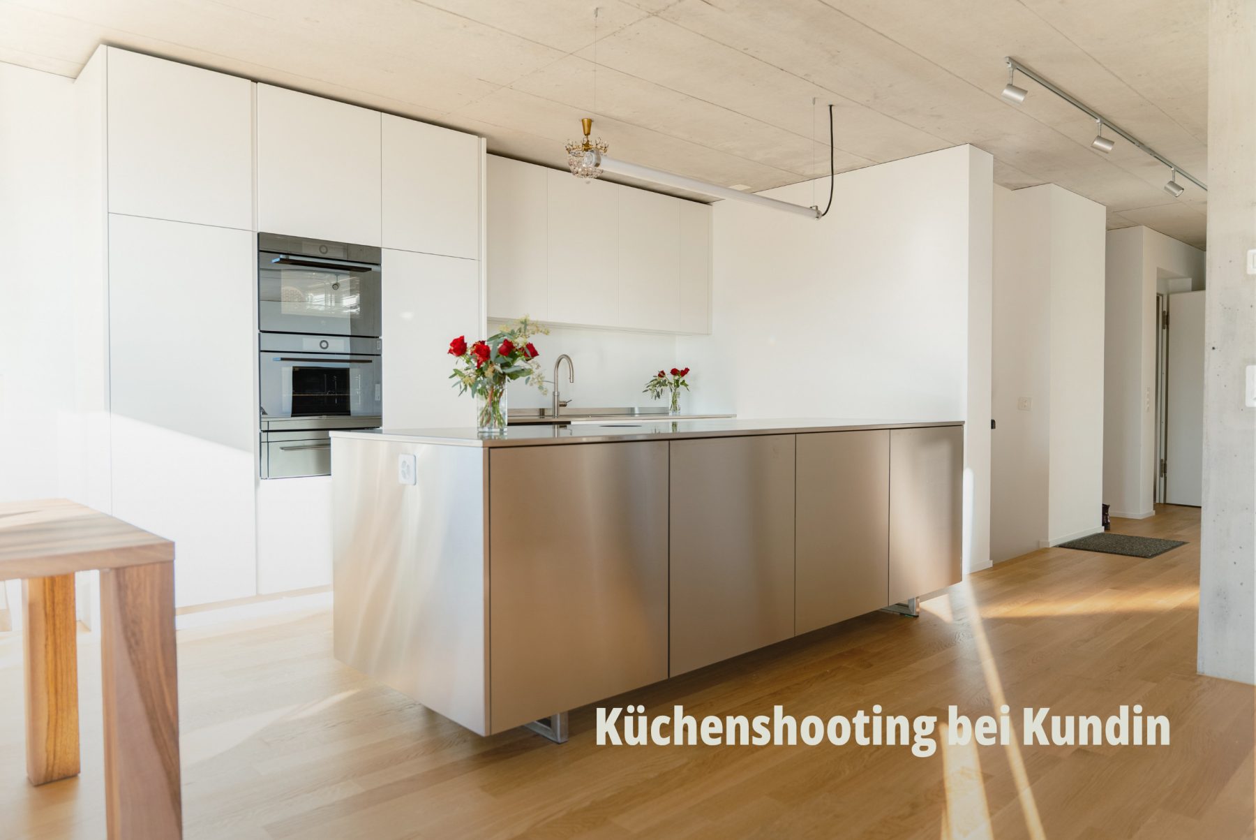 Innenaufnahme einer minimalistischen, weissen Designer Küche mit einer zentralen Kücheninsel aus gebürstetem Metall und hellem Parkettboden; Bard AG. | SR-STUDIOS