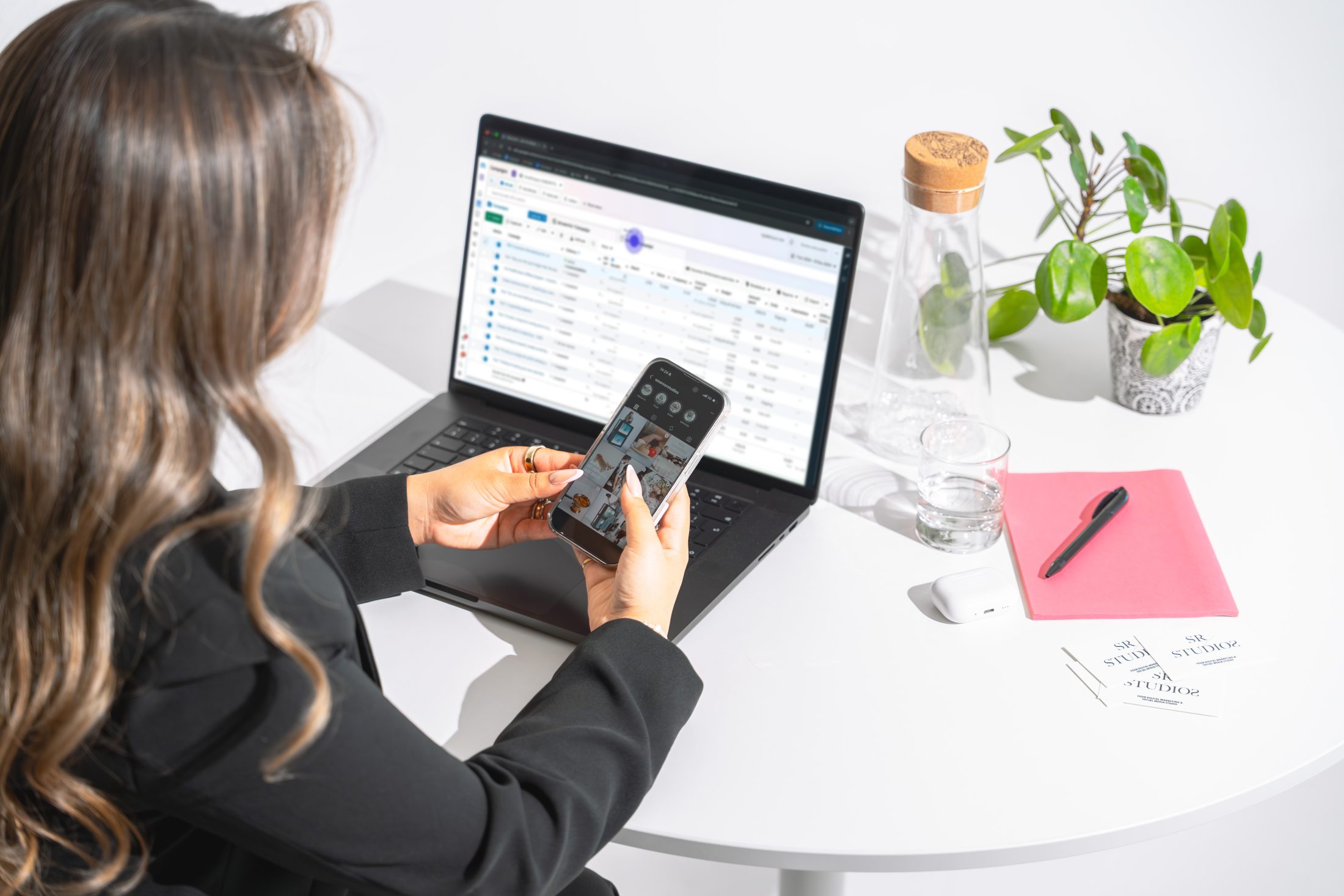 Digital Marketing Specialist ist an ihrem iPhone auf Instagram auf der Seite von SR-STUDIOS; sitzt an einem weissen Tisch mit Laptop, pinkem Notizbuch und Pflanze. | SR-STUDIOS