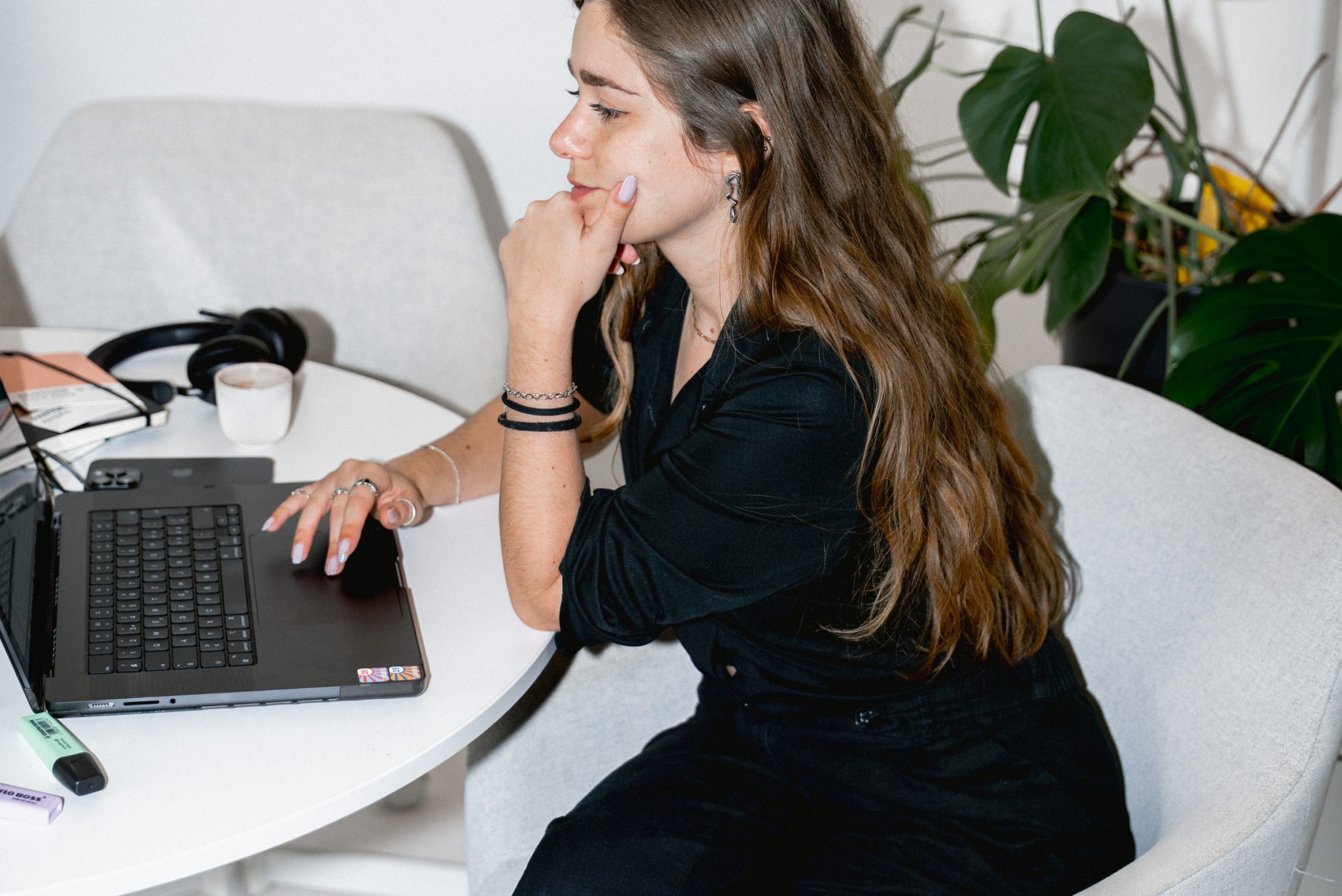 Lora Sibai, Social Media Specialist bei SR-STUDIOS, sitzt in Gedanken versunken an ihrem Laptop an einem weißen runden Tisch, umgeben von einem Kopfhörer und einer Monstera-Pflanze. Sie stützt ihr Kinn nachdenklich auf die linke Hand. | SR-STUDIOS