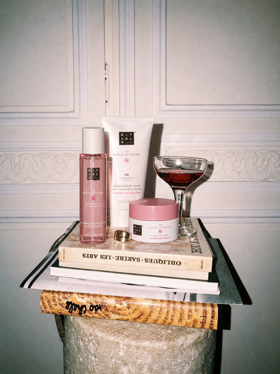 «Eine Chitchat Success Story über Rituals von SR-STUDIOS»: Mehrere «The Ritual of Sakura» Produkte von Rituals, darunter ein Körperspray, eine Körpercreme und ein Körperpeeling, sind stilvoll auf einem Stapel von Büchern arrangiert. Die Bücher liegen auf einer marmorierten Säule. Ein goldenes, gewundenes Schmuckstück und ein Glas mit Rotwein ergänzen das von SR-STUDIOS inszenierte Chitchat-Arrangement. | SR-STUDIOS