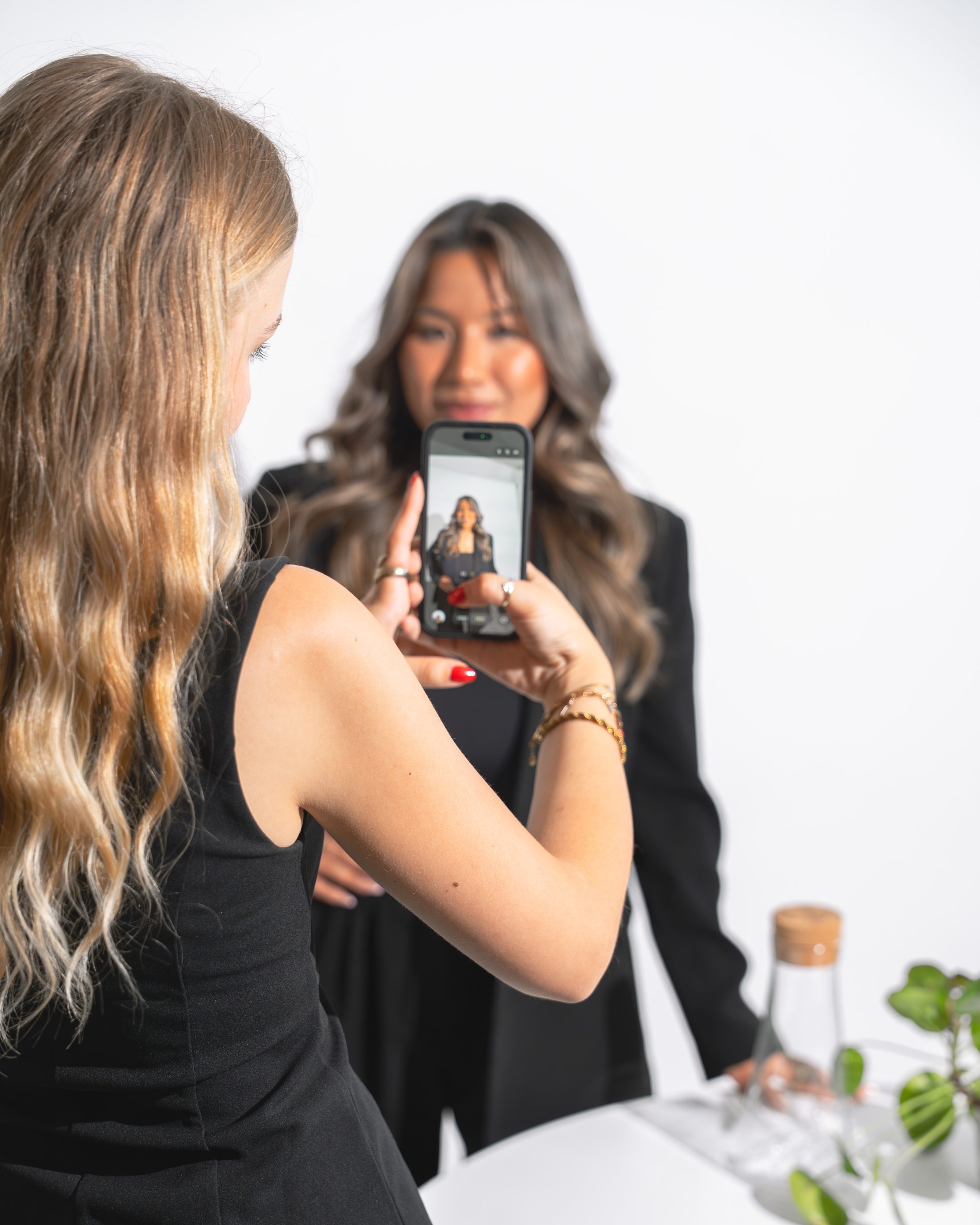 Blonde Frau macht ein Foto mit einem iPhone von einer brünetten Frau die in die Kamera lächelt. Sie stehen vor einem weissen Hintergrund, ein Tisch mit Pflanze ist angeschnitten. Employer Branding by SR-STUDIOS. | SR-STUDIOS