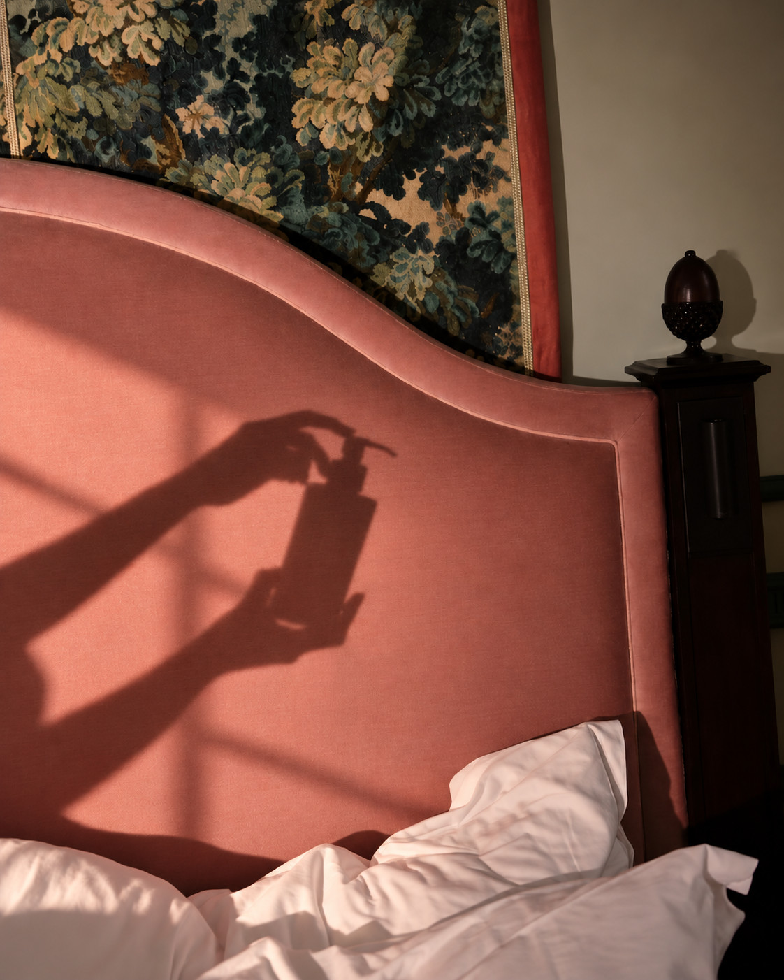 «Eine Chitchat Success Story über Rituals von SR-STUDIOS»: In einem stimmungsvollen Schlafzimmer fällt das Sonnenlicht durch ein Fenster und wirft einen deutlichen Schatten an das rosa Samtkopfteil des Bettes. Der Schatten zeigt zwei Hände, die eine Flasche eines Rituals-Produkts aus dem Chitchat-Shoot von SR-STUDIOS halten. | SR-STUDIOS