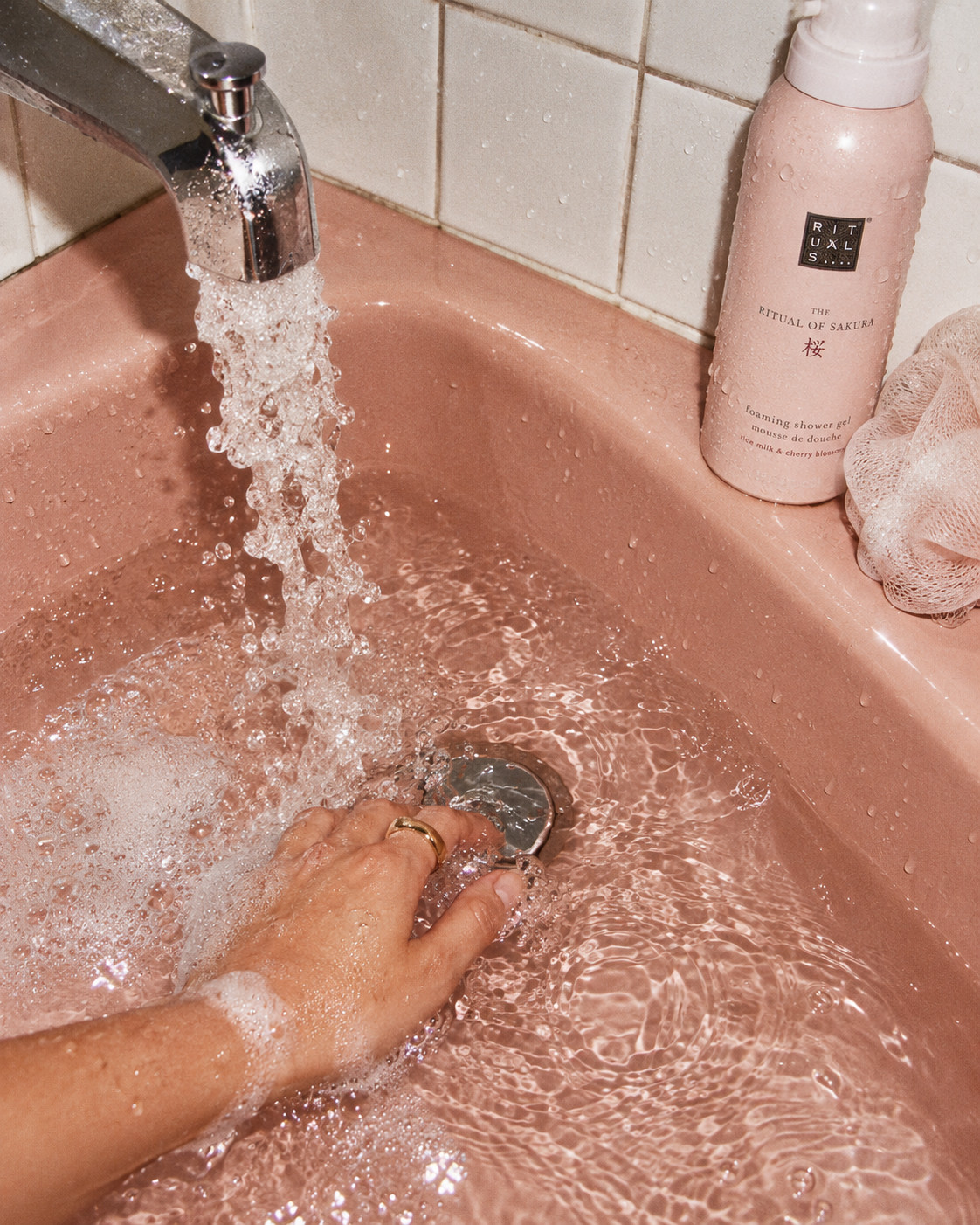 «Eine Chitchat Success Story über Rituals von SR-STUDIOS»: Eine Hand mit einem goldenen Ring taucht in ein rosa Waschbecken, das mit warmem Wasser gefüllt ist. Eine Flasche des «The Ritual of Sakura» Duschschaums von Rituals steht am Rand des Waschbeckens, direkt neben einem weißen Duschschwamm. | SR-STUDIOS