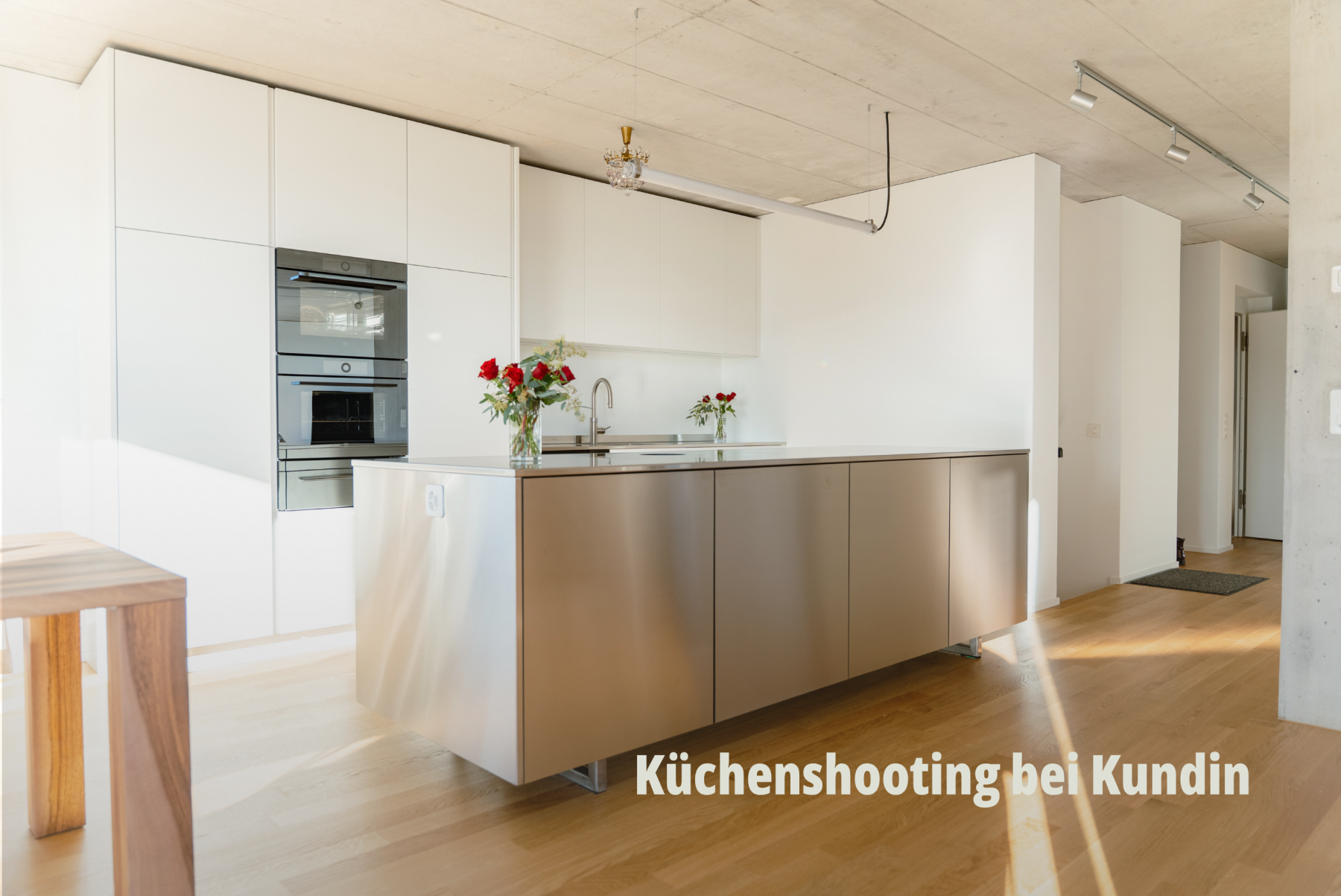 Innenaufnahme einer minimalistischen, weissen Designer Küche mit einer zentralen Kücheninsel aus gebürstetem Metall und hellem Parkettboden; Bard AG. | SR-STUDIOS