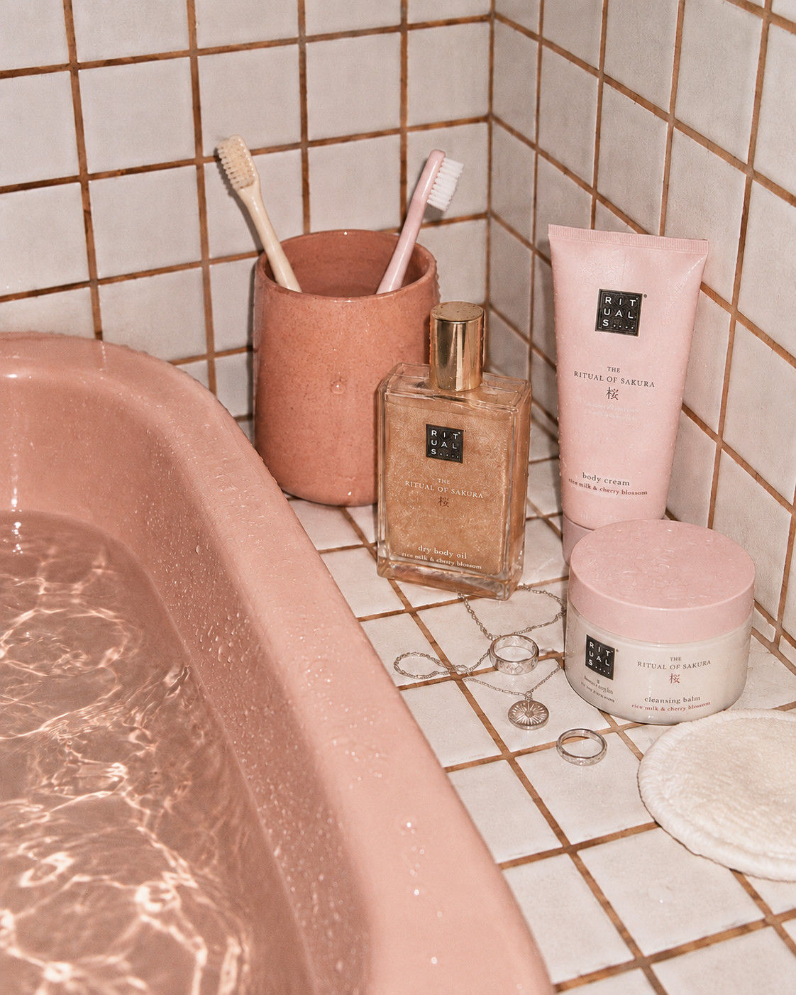 «Eine Chitchat Success Story über Rituals von SR-STUDIOS»: Am Rand einer Badewanne aus rosa Marmor sind verschiedene «The Ritual of Sakura» Produkte von Rituals aufgereiht, darunter ein Reinigungsbalsam, ein Trockenöl und eine Körpercreme. Zwei Zahnbürsten stehen in einem passenden Becher bereit, und ein silbernes Schmuckset liegt auf dem Fliesenrand. | SR-STUDIOS