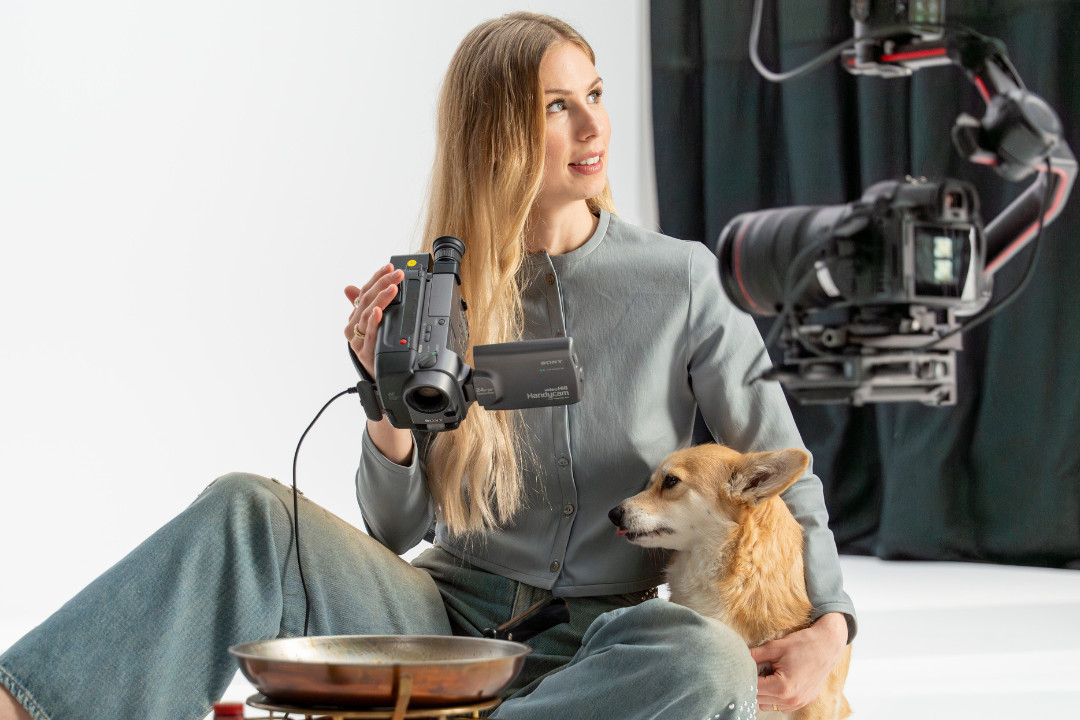 Eine blonde Frau sitzt am Boden des Studio Sets von «SR-STUDIOS has great taste», hält eine alte Videokamera in der Hand und schaut nach oben. Neben ihr sitzt ein Corgi und eine professionelle Studiokamera ist im Vordergrund angeschnitten. | SR-STUDIOS