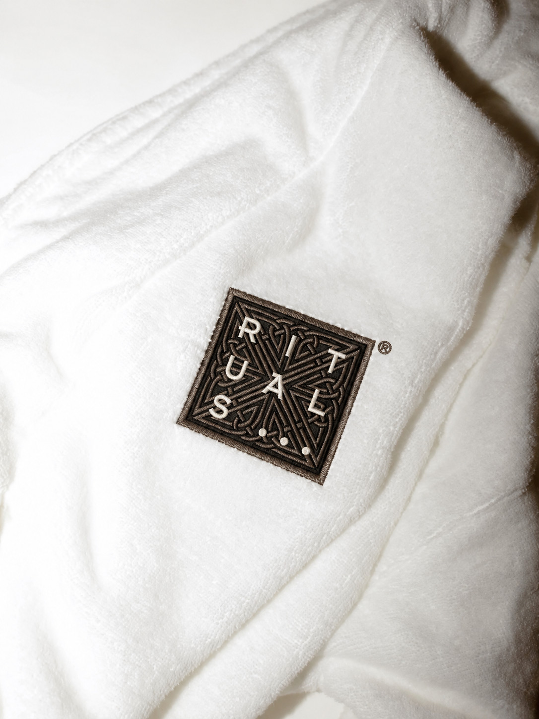«Eine Chitchat Success Story über Rituals von SR-STUDIOS»: Das quadratische Rituals-Logo ist als Stickerei auf dem Ärmel eines flauschigen, weißen Bademantels zu sehen, fotografiert von SR-STUDIOS für Chitchat. Der Bademantel ist weich zusammengelegt. | SR-STUDIOS