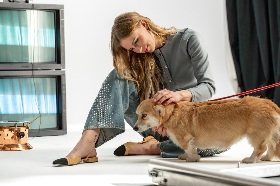 Eine blonde Frau sitzt auf dem Boden eines Studios am Set von «SR-STUDIOS has great taste» und streichelt einen Corgi an der roten Leine. Im Hintergrund stehen zwei alte Röhrenfernseher übereinander. | SR-STUDIOS