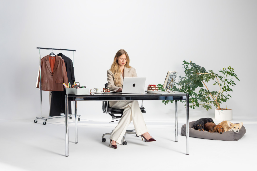 Eine blonde Frau im eleganten hellen Hosenanzug arbeitet an einem Schreibtisch am Laptop in einem hellen Studio. Im Hintergrund stehen ein Kleiderständer mit Jacken und eine Zimmerpflanze. Neben dem Schreibtisch liegen drei kleine Hunde (zwei Dackel und ein Welsh Corgy Pembroke) in einem Hundekorb. Shooting «SR-STUDIOS has great taste». | SR-STUDIOS