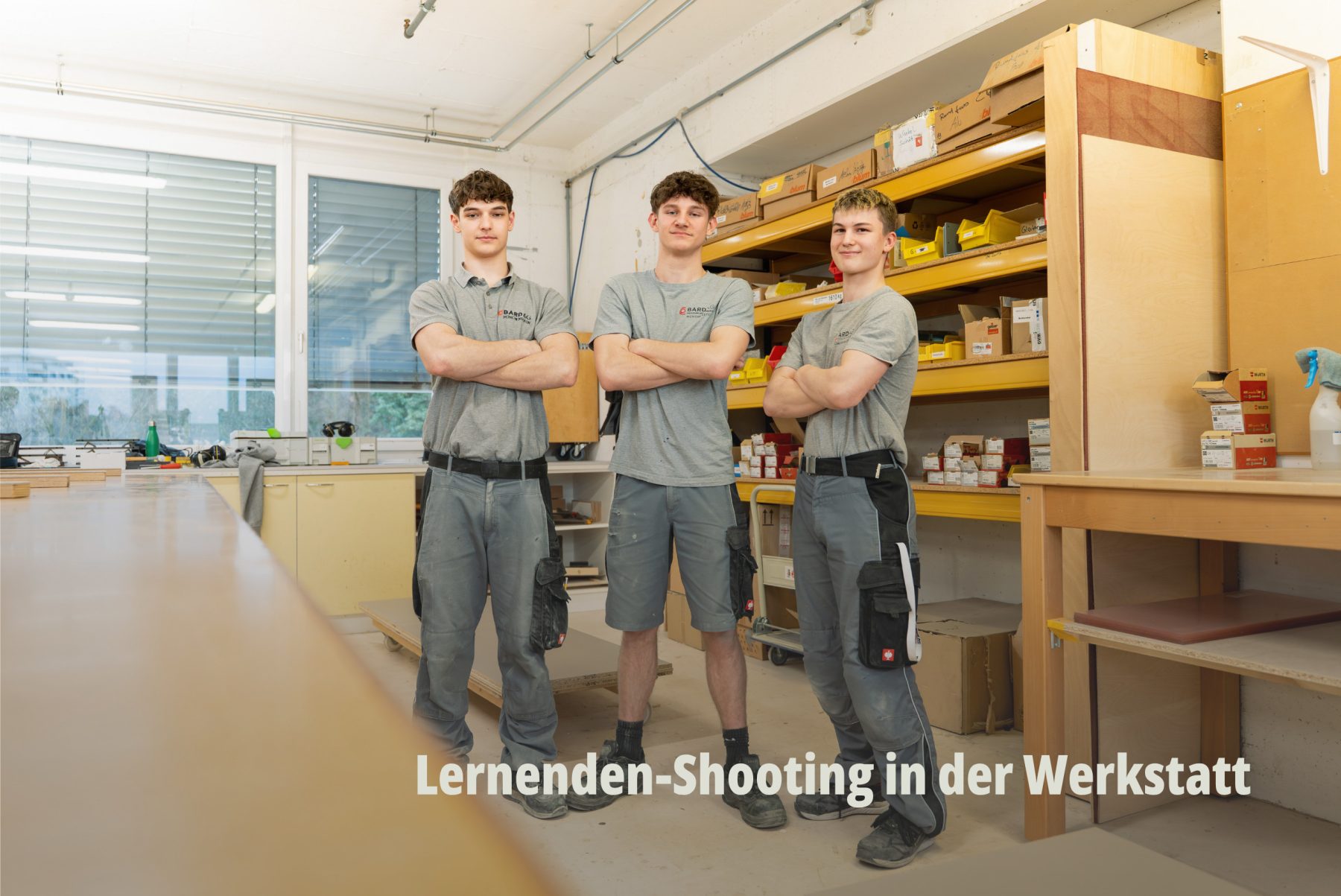 Drei junge Auszubildende / Lernende der Bard AG posieren stolz in ihrer Arbeitskleidung vor einem Materialregal in der Schreinerei Werkstatt. | SR-STUDIOS
