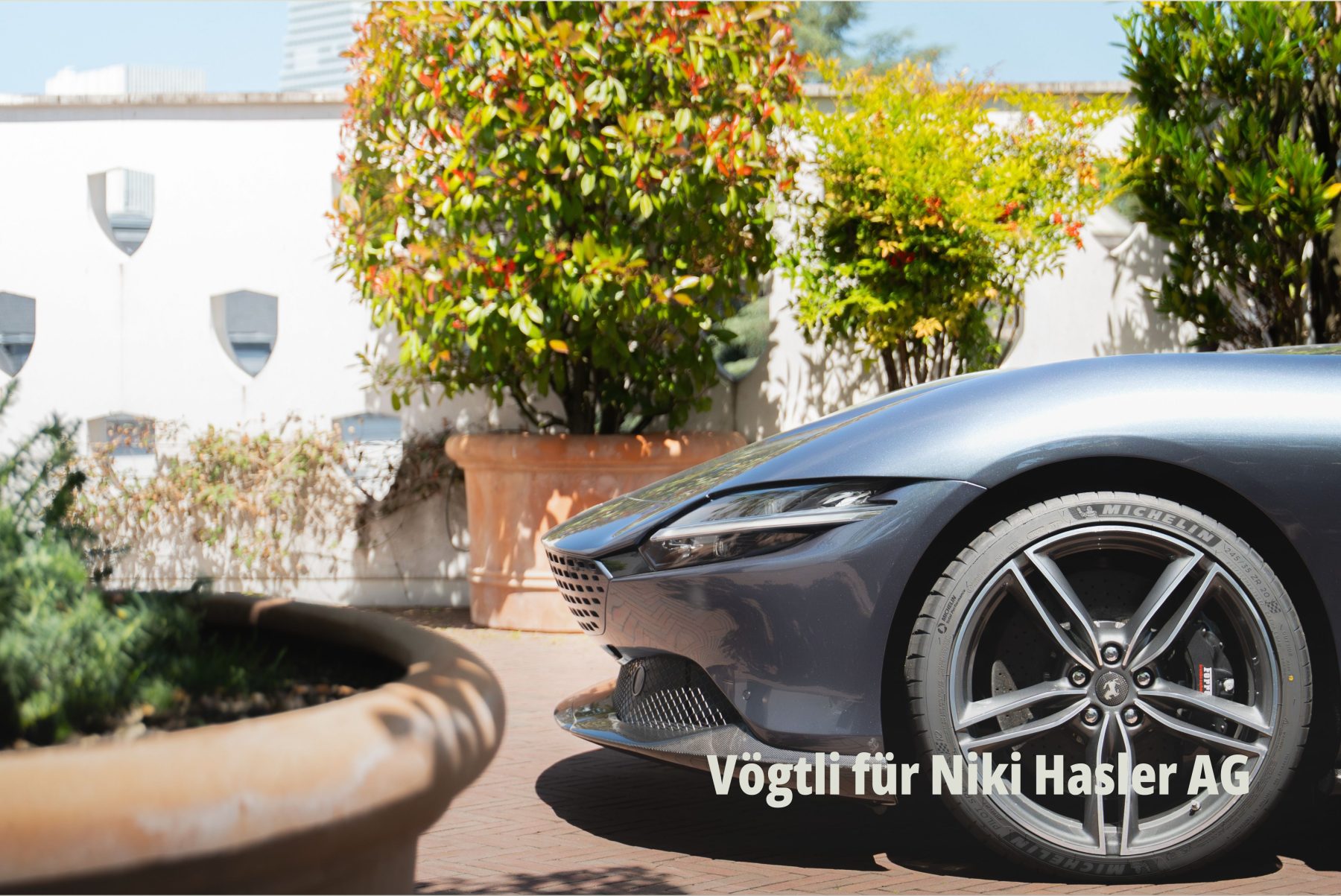 Nahaufnahme der Front und des Vorderrads eines stilvollen, grauen Sportwagens Ferrari mit Michelin Reifen, geparkt auf einem gepflasterten Vorplatz mit üppiger Bepflanzung in großen Terrakottatöpfen. Vögtli Gartenbau GmbH für Niki Hasler AG fotografiert bei SR-STUDIOS. | SR-STUDIOS