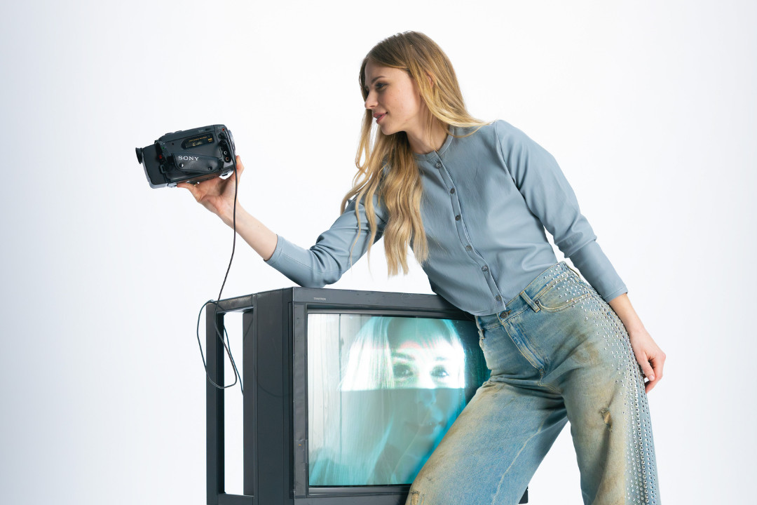 Eine junge Frau in einer blauen Strickjacke und Jeans steht neben einem alten Röhrenfernseher, auf dessen Bildschirm ihr eigenes Gesicht spiegelt. Sie hält eine alte Videokamera in der Hand und blickt darauf. Shooting «SR-STUDIOS has great taste». | SR-STUDIOS