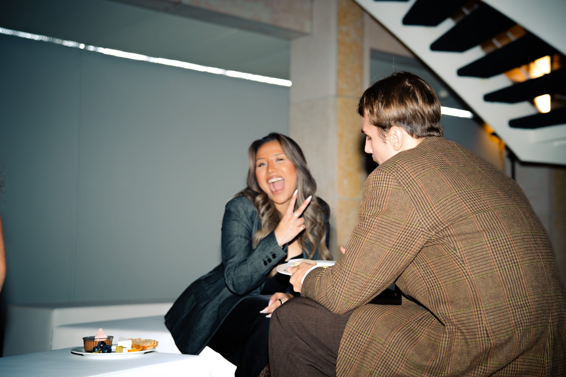 Eine lachende Frau mit dunklen Haaren und ein Mann in einem braunen Tweed-Sakko, sitzen an einem hellen Tisch. Sie schaut in die Kamera, er auf seinen Essensteller, im Hintergrund ist eine Treppe zu sehen. Eventfotografie Embrace the taste by SR-STUDIOS. | SR-STUDIOS