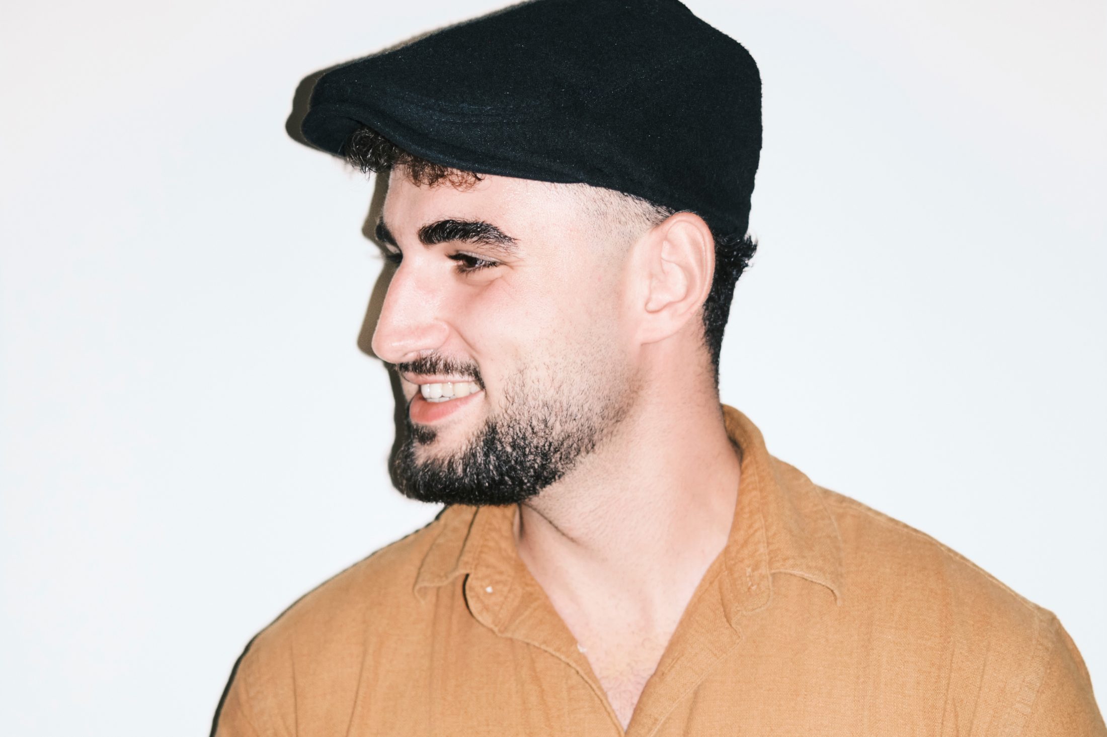 Männlicher Content-Creator mit dunkler Baskenmütze steht lächelnd vor weisser Wand. | SR-STUDIOS
