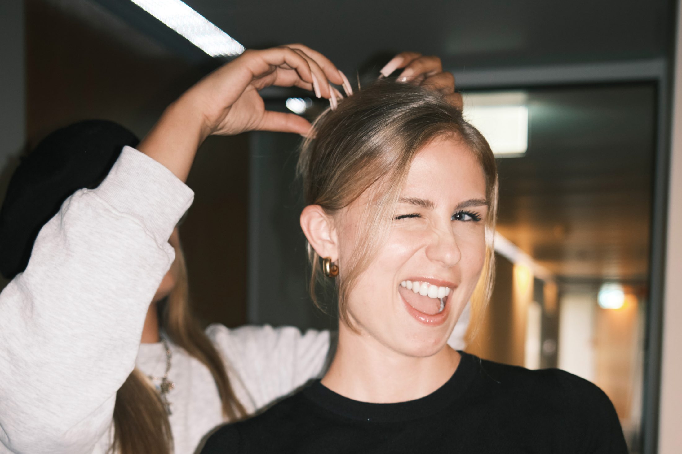 Junge blonde Frau steht lächelnd und zwinkert der Kamera zu, ihre Haare werden gerichtet. | SR-STUDIOS